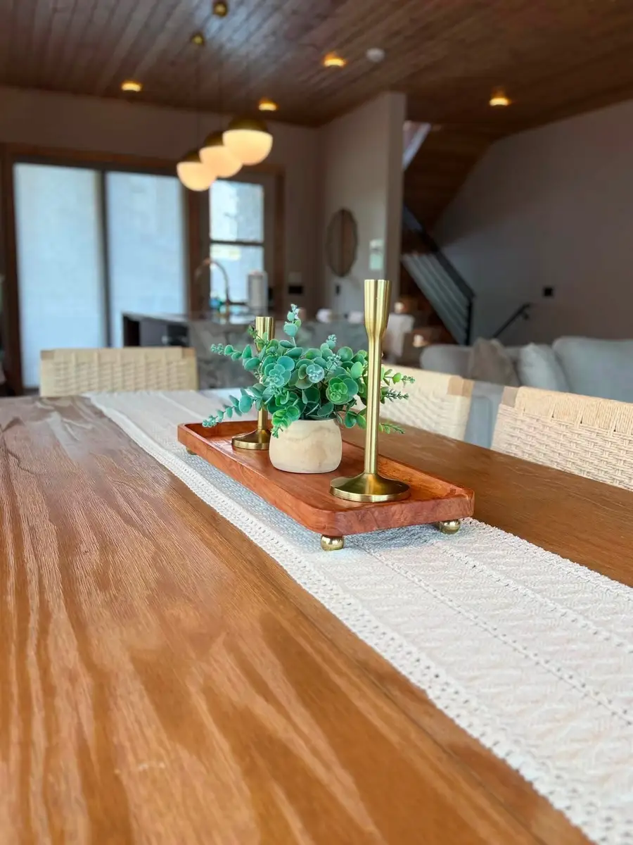 Wood dining table with a runner, brass candlesticks and a small plant, open kitchen behind