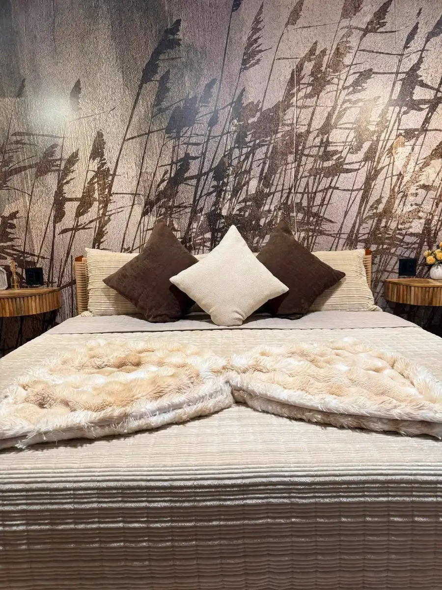 Bed made with cream bedding, brown and white pillows and two faux-fur throws under a reed mural