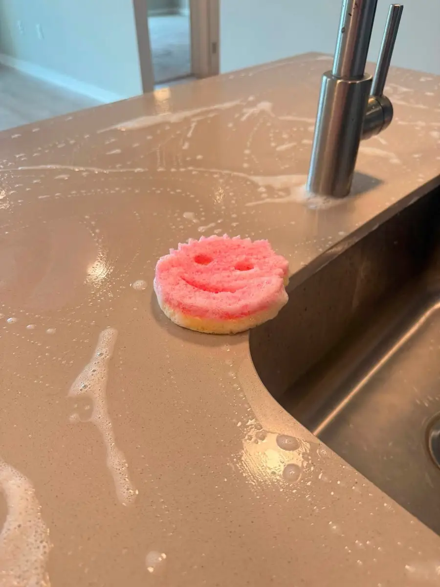 Pink smiley sponge on a soapy quartz counter next to the kitchen sink
